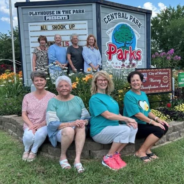 A group of nine people pose in front of a flower bed and sign for Central Park and Playhouse in the Park in Murray, Kentucky, with a "City of Murray Beautification Award" sign also visible.