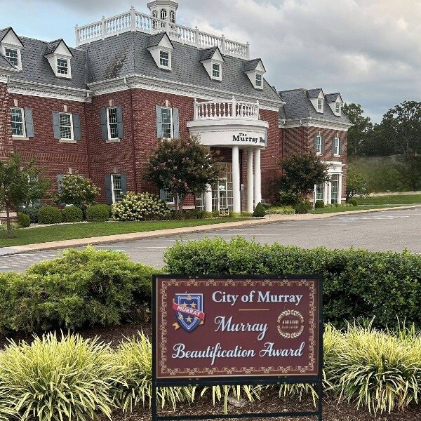 A large red-brick building with white columns and a gray roof stands behind a landscaped lawn. A brown sign in front reads ‘City of Murray, Murray Beautification Award’ with the city’s crest. The scene includes trimmed shrubs, flowers, and a paved driveway under a cloudy sky