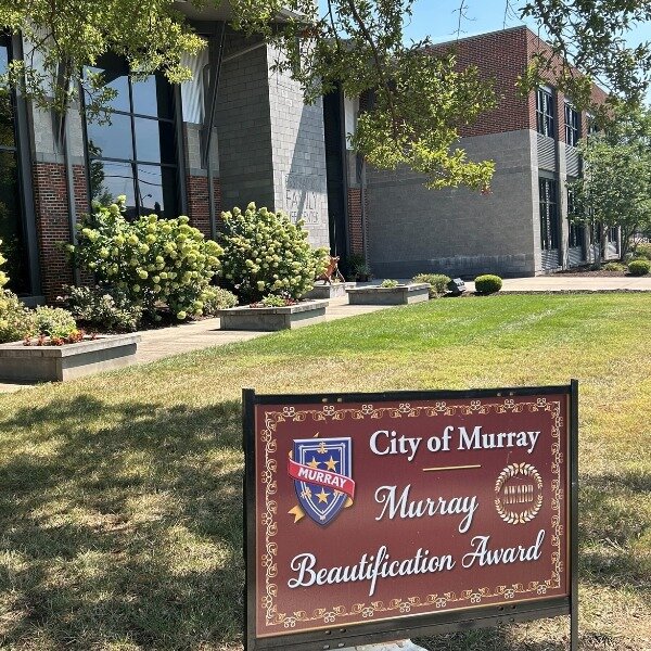 A modern brick and glass building is bordered by green shrubs and flowering plants. In the foreground, a brown sign reads ‘City of Murray, Murray Beautification Award’ with the city crest. Trees cast shade over the grassy area on a sunny day.