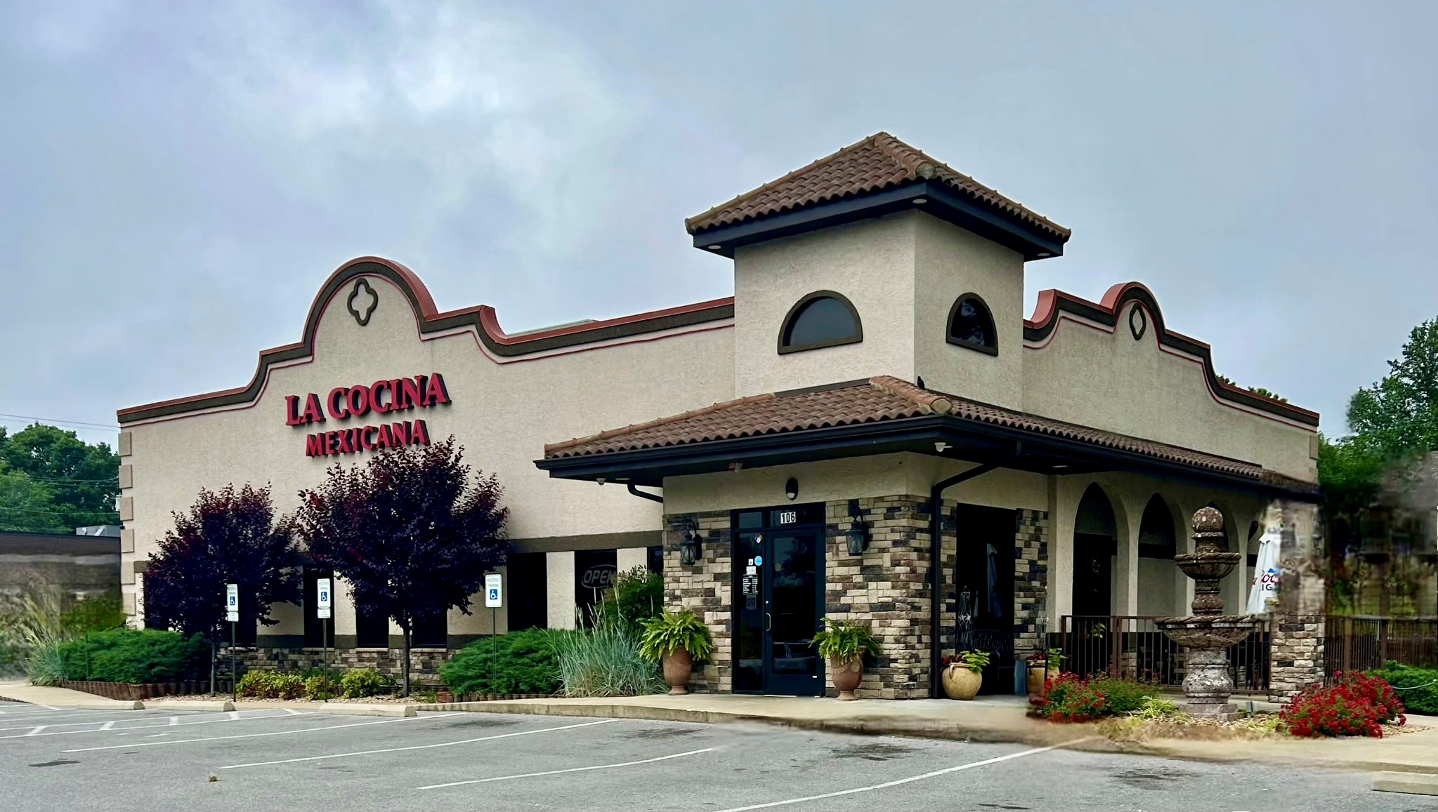 Exterior of La Cocina Mexicana restaurant with a Spanish-style design, including a tiled roof, arched windows, and a stone fountain in front, surrounded by landscaped greenery and an empty parking lot.