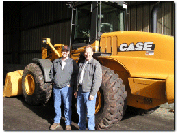 two males with coats on standing in front of a yellow Case heavy duty equipment