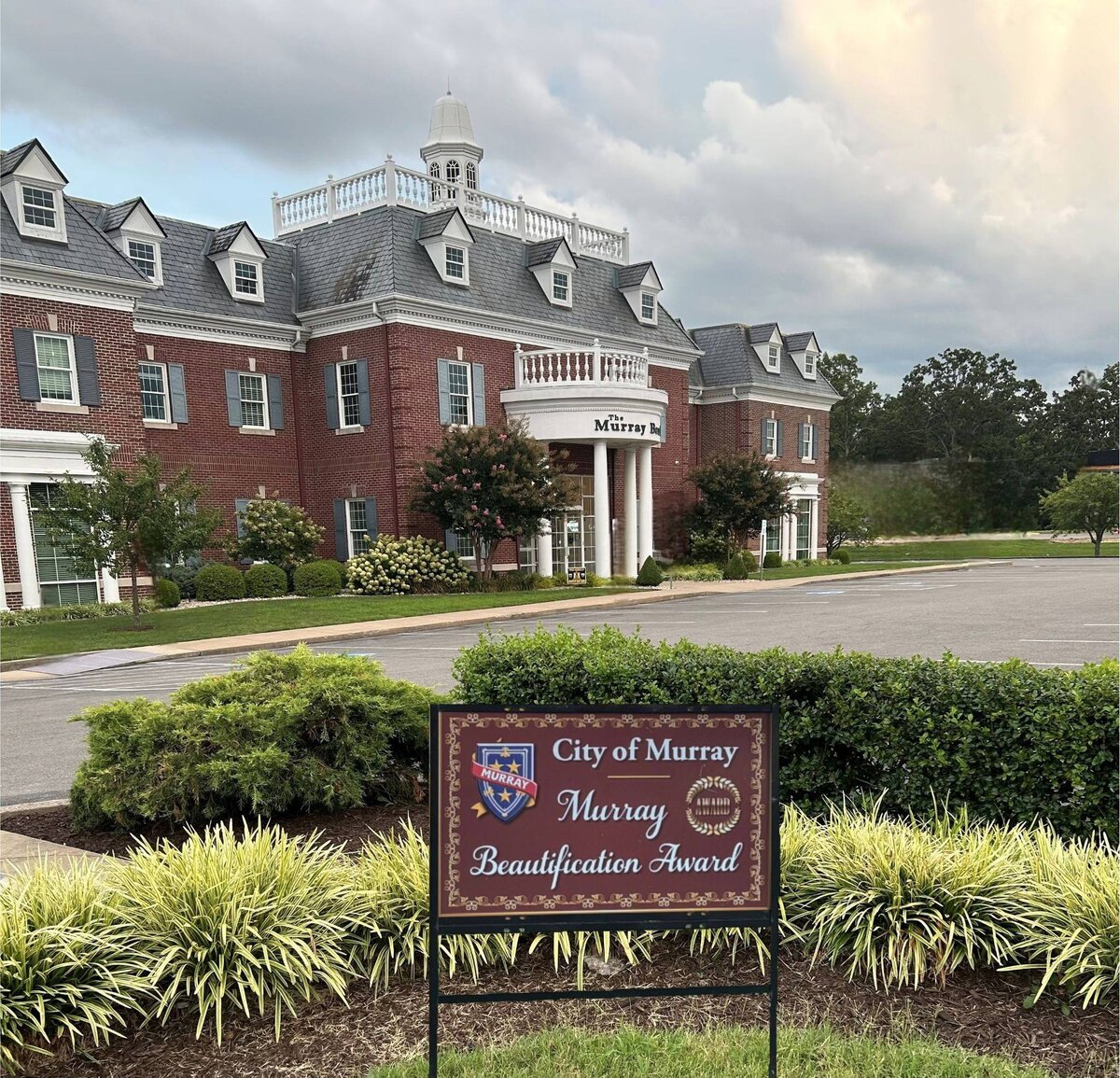 A large red-brick building with white columns and a gray roof stands behind a landscaped lawn. A brown sign in front reads ‘City of Murray, Murray Beautification Award’ with the city’s crest. The scene includes trimmed shrubs, flowers, and a paved driveway under a cloudy sky